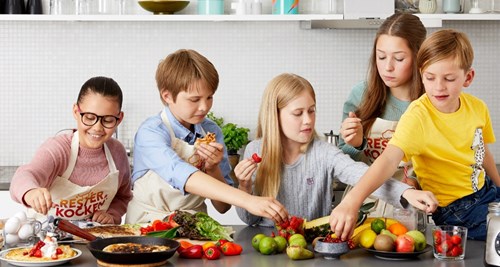 Fem barn i ett kök sträcker sig efter olika matvaror på ett bord.